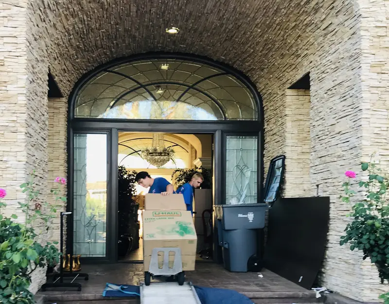 Movers bringing boxed belongings through the arched stone entry of a large home